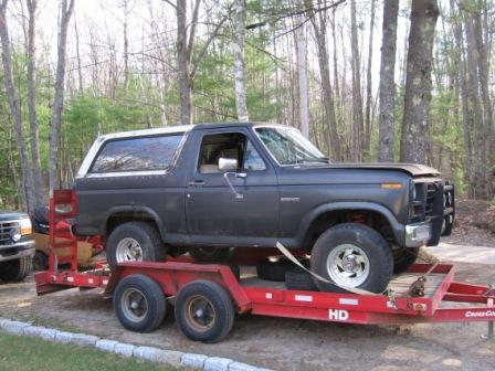 1993 Ford Bronco - A flat-black 1980s Ford Bronco with a white removable hardtop sits chained down on a red tandem-axle equipment trailer in a wooded driveway…