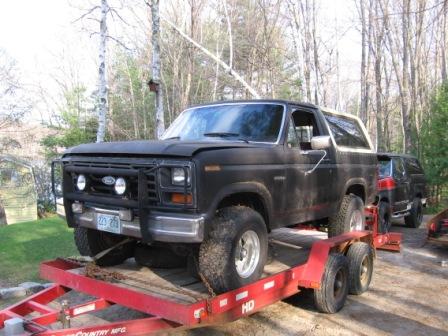 1993 Ford Bronco - Front three-quarter view of a flat-black 1980s Ford Bronco loaded on a red tandem-axle trailer, wearing a tubular brush guard with round au…