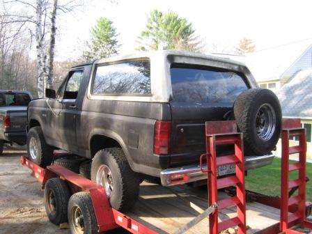 1993 Ford Bronco - Driver-side rear three-quarter view of a flat-black 1980s Ford Bronco chained down on a red equipment trailer, with a weathered white remov…