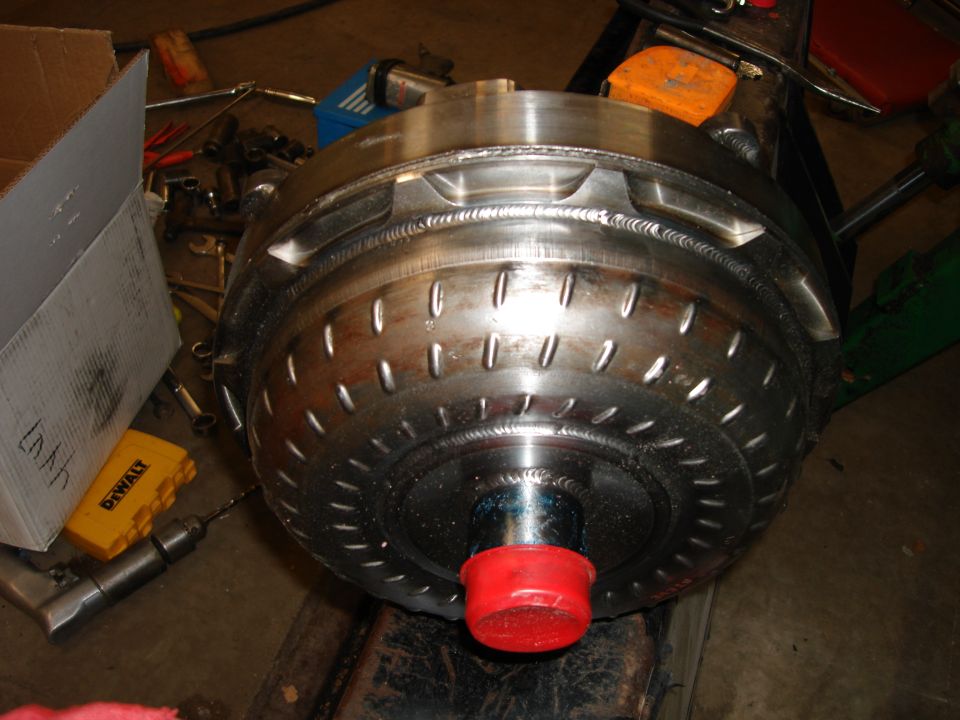 Close-up of a large metallic torque converter with weld marks and indentations, resting on a stand with tools nearby.