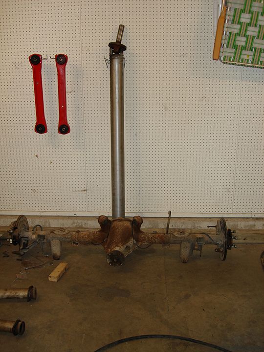 Rusty rear axle housing with driveshaft upright on shop floor below red control arms