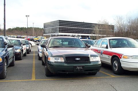 2001 Ford Crown Victoria - Several police cruisers are parked in a lot, with a modern office building in the background.