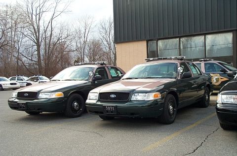 2001 Ford Crown Victoria - Several police vehicles are parked in a lot outside a building with large windows.