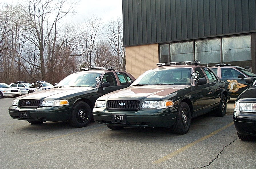 Two dark green police cars parked in front of a building, one in the foreground with its grille and headlights visible