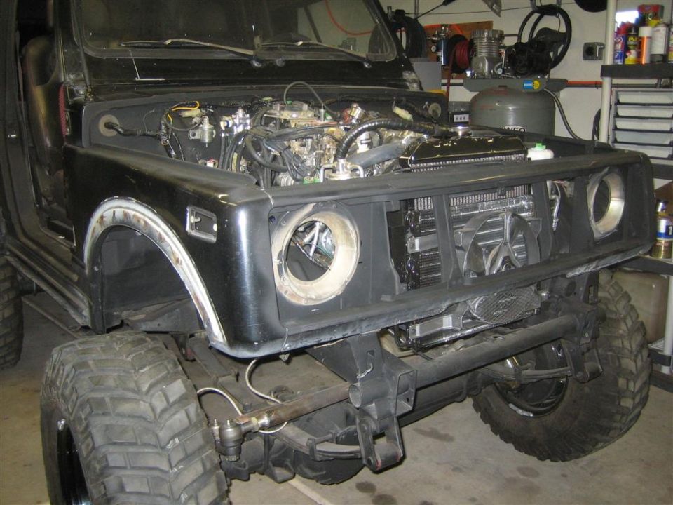 Front view of a black Suzuki Samurai with hood open, showing engine and radiator, modified tires.