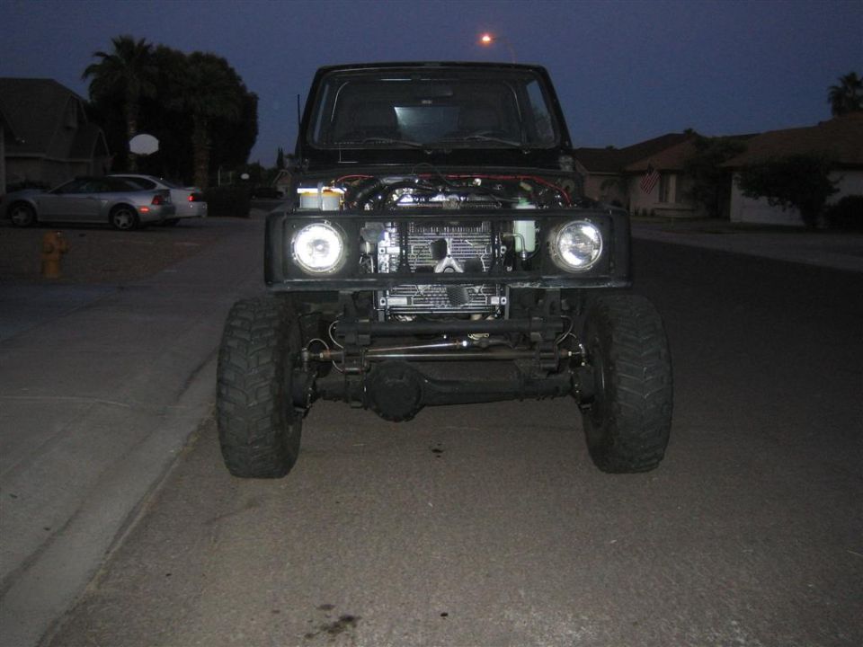 Modified black Suzuki Samurai with hood open, front view, parked on street at dusk.
