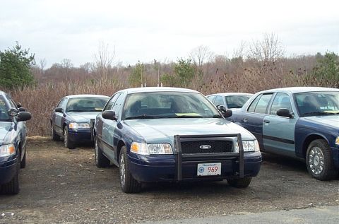 2001 Ford Crown Victoria - Multiple Ford Crown Victoria police cars are parked in a gravel lot with dry brush in the background.