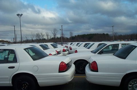 2001 Ford Crown Victoria - A large group of white sedans, likely police or taxi vehicles, are parked in an outdoor lot under a cloudy sky.