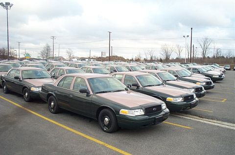 2001 Ford Crown Victoria - A row of green-and-tan two-tone Ford Crown Victoria police sedans lined up nose-out in a paved lot under an overcast sky, all wearing black…