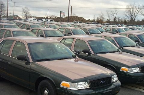 2001 Ford Crown Victoria - Rows of identical two-tone green over tan Ford Crown Victoria Police Interceptor sedans lined up in a fleet storage lot, each with black st…