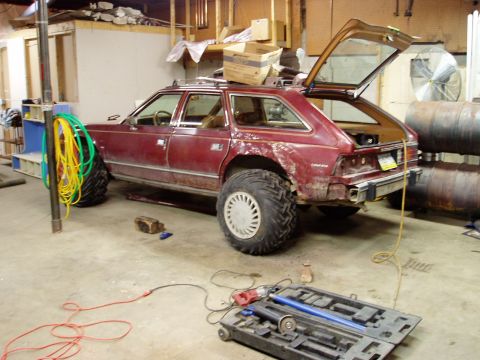2000 Ford E350 - A modified maroon station wagon with oversized tires is parked in a garage. The rear hatch is open, revealing the interior, and tools are l…