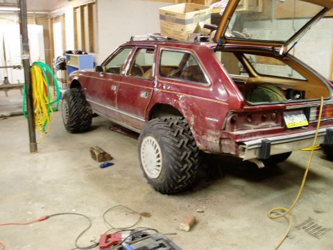 2000 Ford E350 - A modified maroon station wagon with oversized off-road tires is parked in a garage. The rear liftgate is open, revealing the interior and…