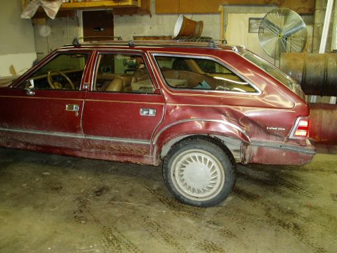 2000 Ford E350 - The side view of a damaged, dark red station wagon parked indoors on a dirty concrete floor. The rear passenger side of the vehicle shows s…