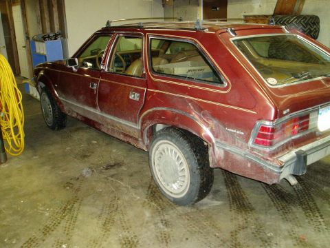 2000 Ford E350 - A dusty, maroon AMC Eagle wagon is parked indoors on a concrete floor with tire tracks. The rear passenger side of the vehicle is visible,…
