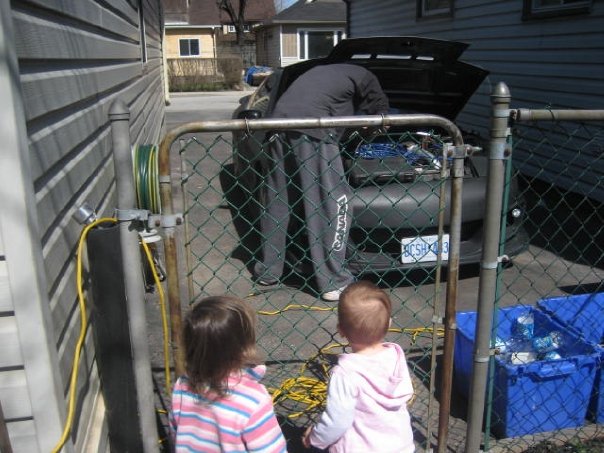 Person working on car engine, viewed through fence, with two children in foreground.