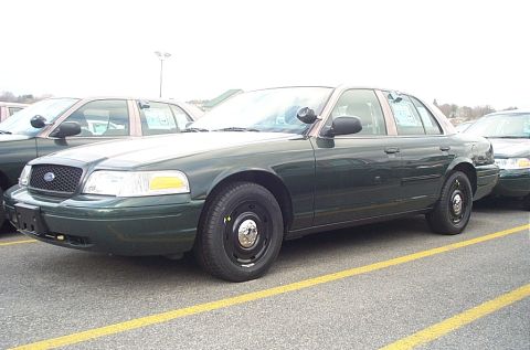 2001 Ford Crown Victoria - Two dark green Ford Crown Victoria police cars are parked side-by-side in a lot. One has a push bumper.