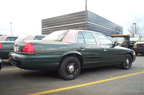 2001 Ford Crown Victoria - A dark green police car is parked in a lot, with another similar vehicle to its left and a yellow truck to its right.