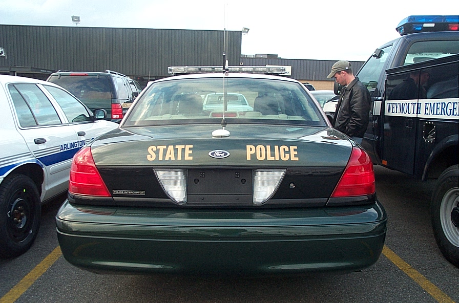 Rear of a dark green Ford Crown Victoria police car with 'STATE POLICE' on the trunk. Another police car and a van are nearby