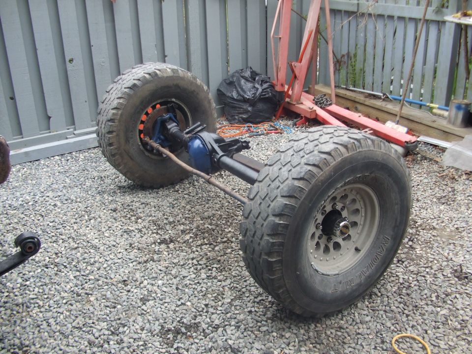 Bare rear axle with blue differential and two large off-road tires resting on gravel