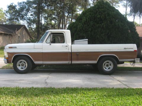1972 Ford F-100 - The side profile of a two-tone white and brown 1972 Ford F-100 pickup truck parked on a paved surface in front of a house and trees.