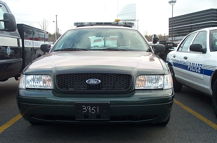 Front view of a dark green Ford Crown Victoria police car with a light bar. A white police car is to its right. License plate reads 3951