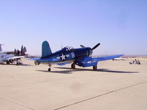 1990 Ford Bronco - A vintage blue F4U Corsair fighter plane is parked on an airfield under a clear blue sky. Another vintage aircraft is parked to the left of…