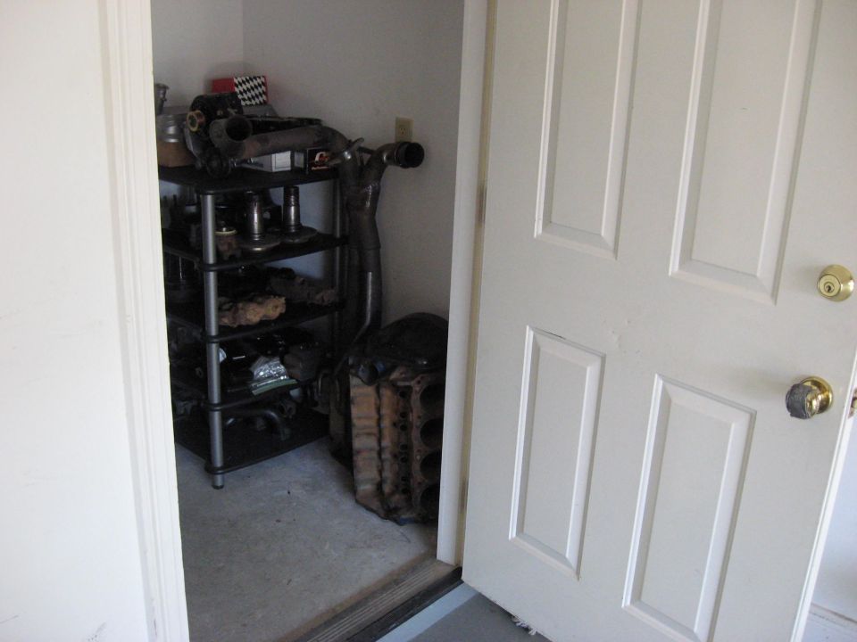 Car parts on shelves and an engine block in a storage closet next to a white door.