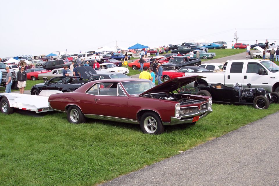 Maroon Pontiac coupe with hood open at a car show, parked next to a white trailer.