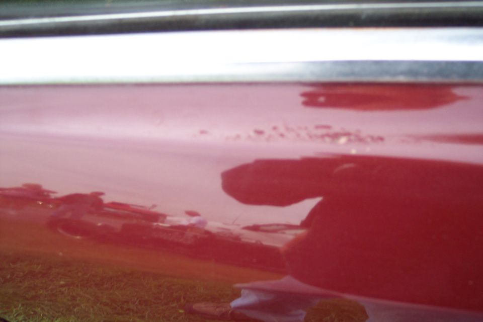 Close-up of a red car's side panel with reflections and minor paint damage.