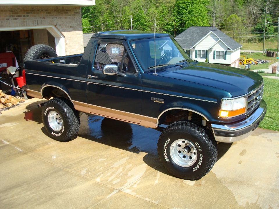 Dark green Ford Bronco with tan trim, lifted suspension, and off-road tires parked on a wet driveway