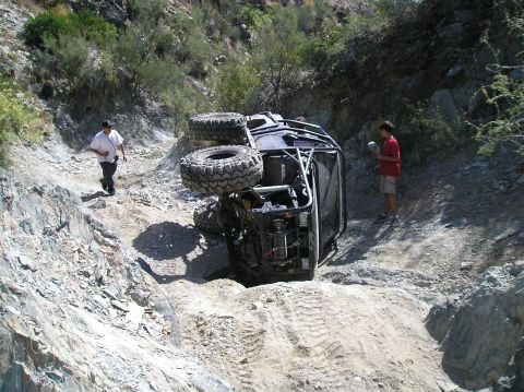 1986 Toyota Hilux - An off-road vehicle has overturned on a steep, rocky trail, with two individuals observing the scene.