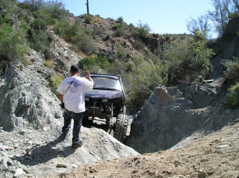 1986 Toyota Hilux - A person stands by a heavily modified off-road vehicle as it navigates a challenging, rocky terrain.