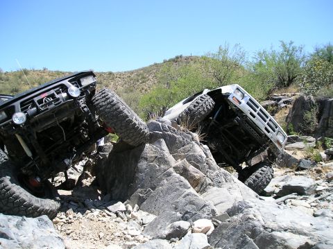 1986 Toyota Hilux - Two modified trucks navigate a challenging rocky terrain on a sunny day.