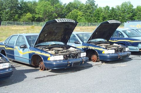 2001 Ford Crown Victoria - Two blue police cars, likely decommissioned, sit with hoods open and missing wheels.