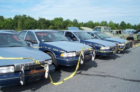 2001 Ford Crown Victoria - Several police cars are parked in a row, some marked with yellow caution tape, under a partly cloudy sky.