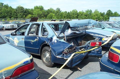 2001 Ford Crown Victoria - A blue-and-gray two-tone state police sedan sits in a salvage lot with its entire rear end crushed and torn open, trunk lid buckled upward…