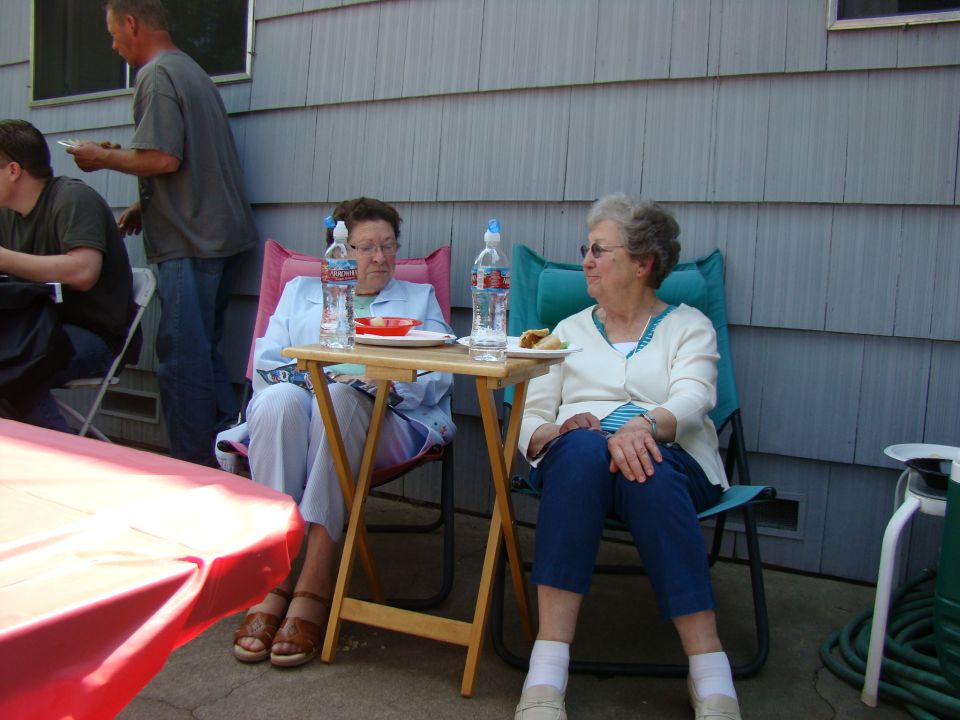 Two older women sit at a folding table outside, with a man standing behind them. Water bottles and plates of food are on the table