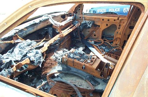 2001 Ford Crown Victoria - The interior of this vehicle is destroyed by fire, with only charred remains visible. A state police vehicle is seen outside.