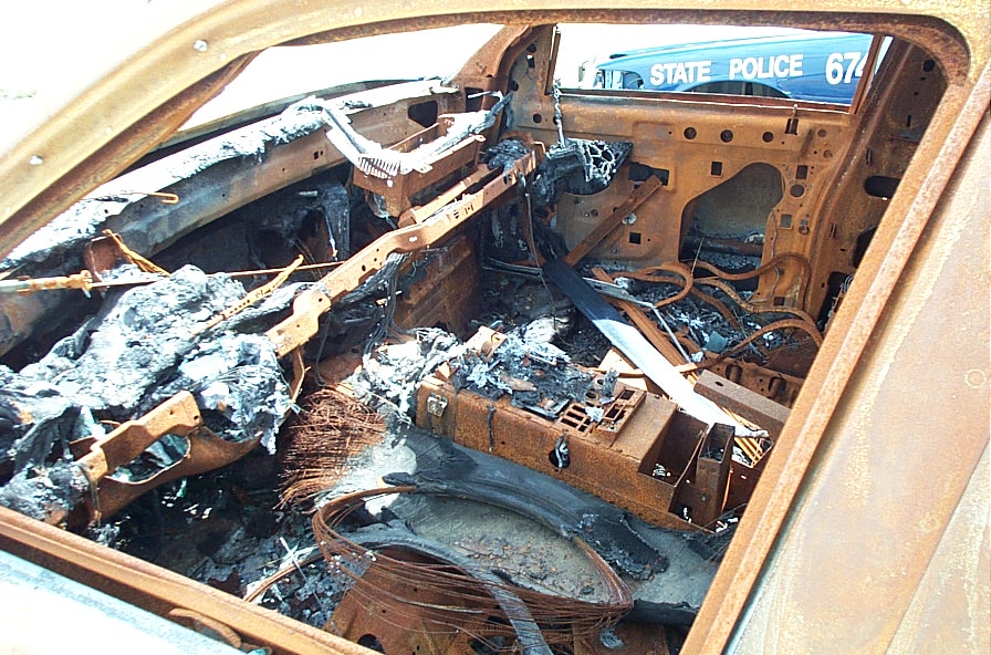 Interior of a completely burnt-out car, showing melted dashboard and seats. A state police vehicle is visible through the window