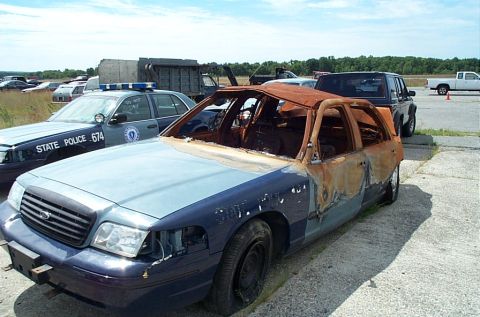 2001 Ford Crown Victoria - Burned-out blue and grey Ford Crown Victoria police sedan sitting on cracked concrete, its roof, doors and rear quarter scorched to bare ru…