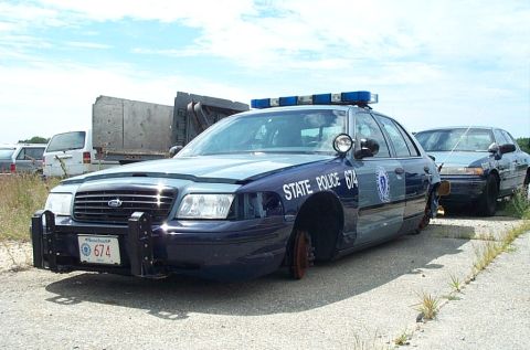 2001 Ford Crown Victoria - A dark blue Ford Crown Victoria Massachusetts State Police cruiser (unit 674) sits abandoned in a gravel lot with its front wheels missing,…