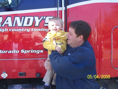 1972 Ford Ranchero - A man holds a child in front of a large red truck with "Randy's High Country Towing" written on its side.