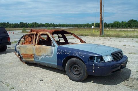 2001 Ford Crown Victoria - Passenger-side view of a fire-gutted Ford Crown Victoria police sedan on a weedy concrete pad, with faded "STATE POLICE" lettering still vi…