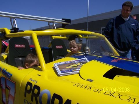 1972 Ford Ranchero - A yellow race car with a roll cage and racing seats is shown with two children inside and a man standing behind it.
