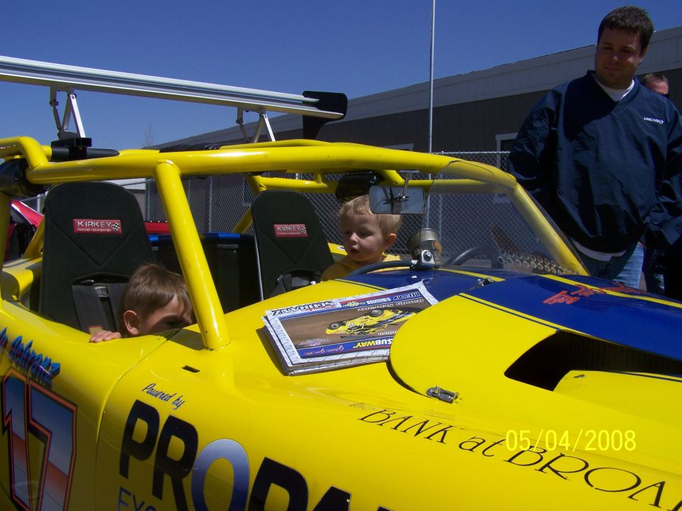 Yellow race car with blue accents, roll cage, and racing seats. Two young boys are in the driver and passenger seats