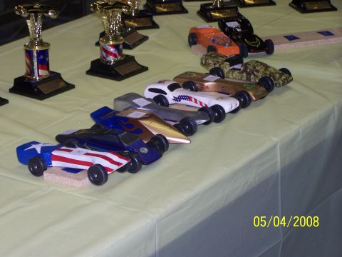 1972 Ford Ranchero - Several custom-painted pinewood derby cars are displayed on a table, ready for competition, alongside trophies.