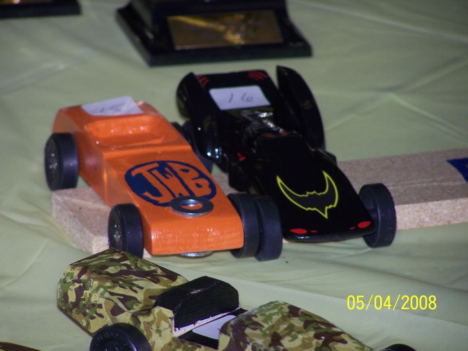 Two pinewood derby cars, one orange with JWB logo, one black with bat symbol, on a table