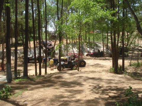1989 Ford Bronco II - A group of off-road vehicles are parked in a clearing surrounded by trees, with several people milling about.