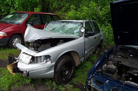 2001 Ford Crown Victoria - A damaged 2001 Ford Crown Victoria sits in a yard with other vehicles, showing extensive front-end damage and a cracked windshield.
