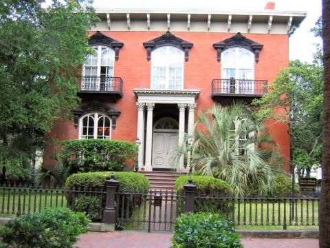1985 Ford LTD Crown Victoria - A grand historic brick house with arched windows and decorative balconies stands behind a black wrought-iron fence, with lush trees and shr…